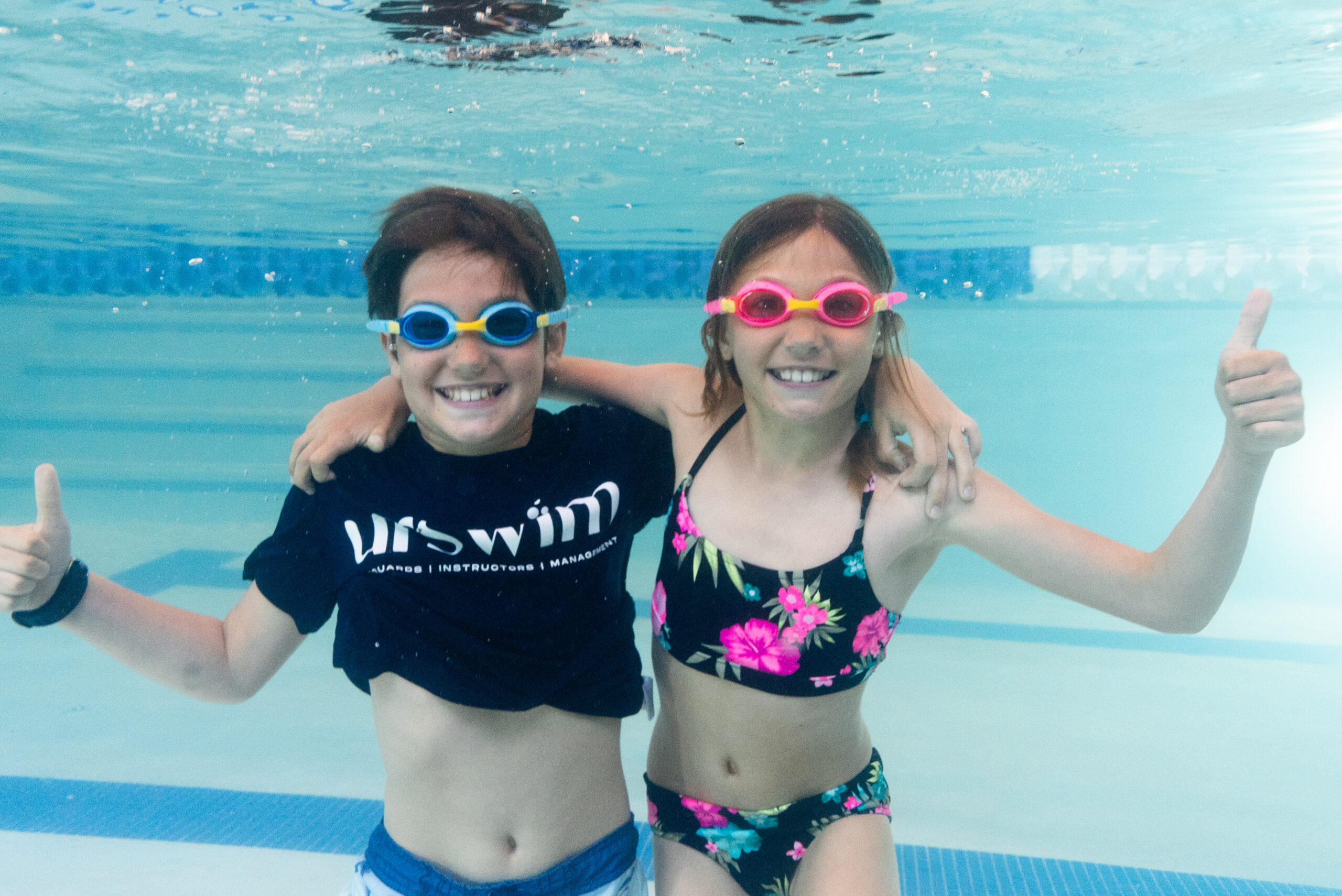 Two children smiling underwater in a swimming pool, enjoying their swim class with excitement and teamwork