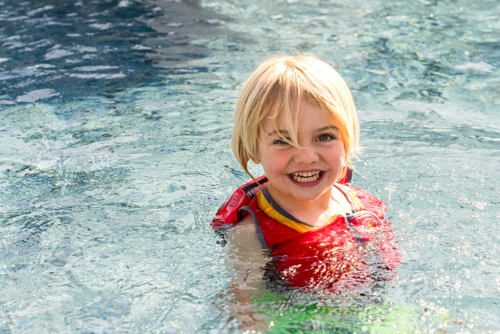 Young child smiling in the pool wearing a red life jacket during swim practice